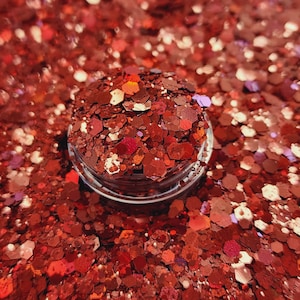 May include: A close-up of a small jar filled with red, orange, and gold glitter. The glitter is loose and scattered, creating a sparkly and festive effect.