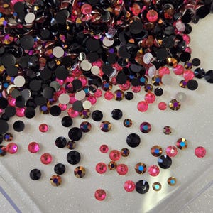 Puede incluir: Primer plano de una variedad de pequeños diamantes de imitación redondos e iridiscentes en colores negro, rosa y arcoíris. Los diamantes de imitación están esparcidos sobre una superficie blanca.