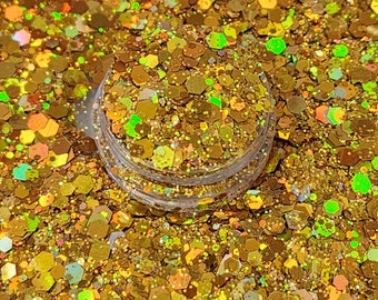 Puede incluir: Un recipiente de plástico transparente lleno de purpurina dorada. La purpurina es una mezcla de formas hexagonales de varios tamaños, con reflejos iridiscentes y verdes. La purpurina está esparcida alrededor del recipiente, creando un efecto brillante.