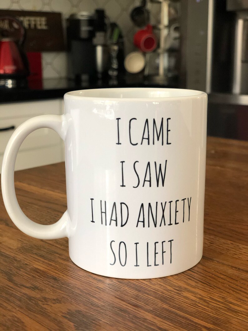 May include: White ceramic mug with the text "I CAME I SAW I HAD ANXIETY SO I LEFT" in black. The mug has a curved handle and is on a wooden surface. The background is blurred, showing kitchen items.