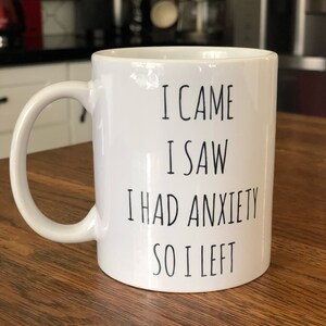 May include: White ceramic mug with a curved handle. The mug has black text that reads "I CAME I SAW I HAD ANXIETY SO I LEFT". The mug is on a wooden surface.