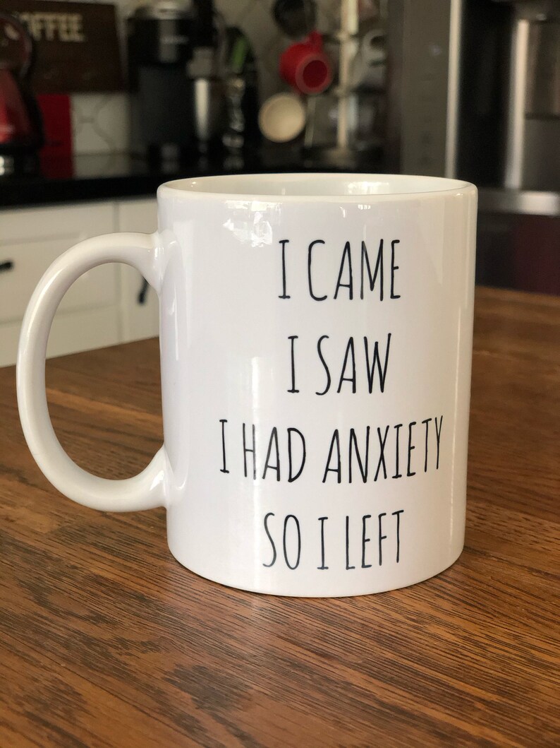May include: White ceramic mug with a curved handle. The mug has black text that reads "I CAME I SAW I HAD ANXIETY SO I LEFT". The mug is on a wooden surface.