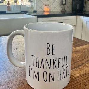 May include: White ceramic mug with a curved handle. The mug features black text that reads "BE THANKFUL I'M ON HRT!". The mug is sitting on a wooden surface.