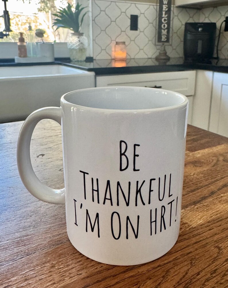 May include: White ceramic mug with a curved handle. The mug features black text that reads "BE THANKFUL I'M ON HRT!". The mug is sitting on a wooden surface.