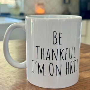 May include: White ceramic mug with a curved handle. The mug features black text that reads "BE THANKFUL I'M ON HRT!". The mug is on a wooden surface.