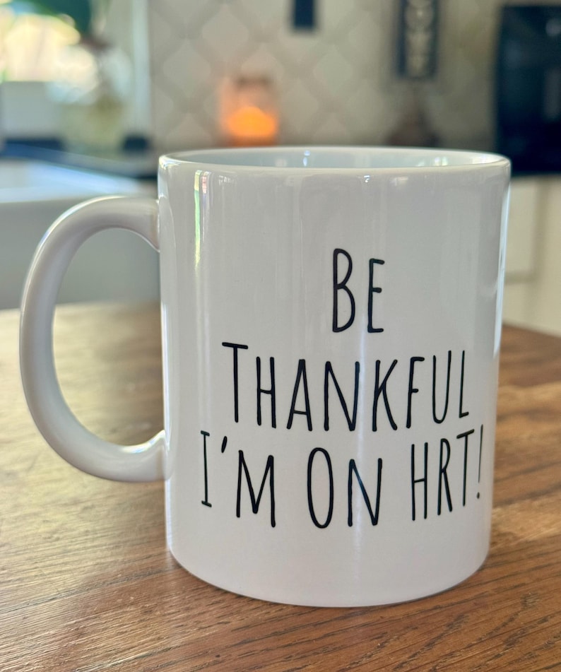 May include: White ceramic mug with a curved handle. The mug features black text that reads "BE THANKFUL I'M ON HRT!". The mug is on a wooden surface.