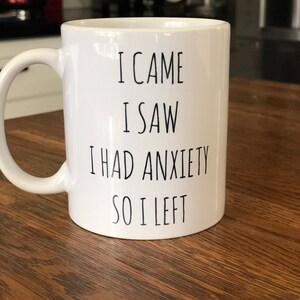 May include: White ceramic mug with a curved handle. The mug has black text that reads "I CAME I SAW I HAD ANXIETY SO I LEFT". The mug is on a wooden surface.