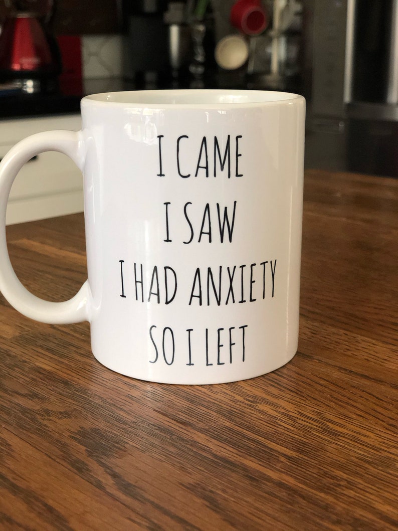 May include: White ceramic mug with a curved handle. The mug has black text that reads "I CAME I SAW I HAD ANXIETY SO I LEFT". The mug is on a wooden surface.