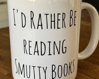 May include: White ceramic mug with a black handle. The mug features the text "I'D RATHER BE READING SMUTTY BOOKS" in a black, handwritten-style font. The mug is sitting on a wooden surface.