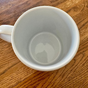 May include: A pale blue ceramic mug with a white handle. The mug is empty and rests on a wooden surface. The interior of the mug is a lighter shade of blue.