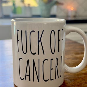 May include: White ceramic mug with a curved handle. The mug features the text "FUCK OFF CANCER!" in black, bold, uppercase letters. The mug is sitting on a wooden surface.