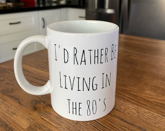 May include: White ceramic mug with a curved handle. The mug features black text that reads "I'D RATHER BE LIVING IN THE 80'S". The mug is sitting on a wooden surface.