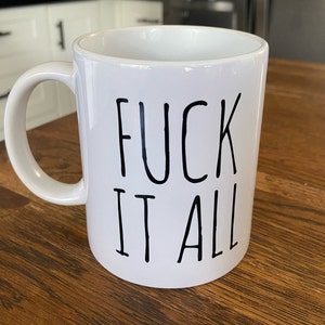 May include: White ceramic mug with the phrase "FUCK IT ALL" in black, handwritten-style lettering. The mug has a curved handle and is set against a wooden surface.