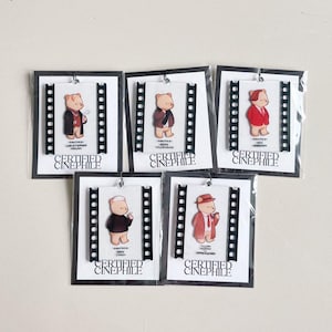 May include: Five bear-shaped acrylic charms, each dressed in different outfits, are displayed on white cards with black borders. The charms are set against a film strip design. The text "CERTIFIED CINEPHILE" is printed on each card.