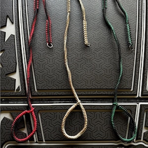May include: Three braided paracord lanyards with metal carabiners at the top and circular loops at the bottom. The lanyards feature color combinations of red and black, solid tan, and green and black. Each cord is woven in a tight, textured pattern.