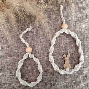 Könnte beinhalten: Zwei handgefertigte Makramee-Osterornamente. Eines ist eine einfache Schlaufe mit einer Holzperle, das andere zeigt einen Holzhasen mit einem flauschigen weißen Schwanz. Beide sind aus cremefarbenem, gedrehtem Kordel gefertigt.