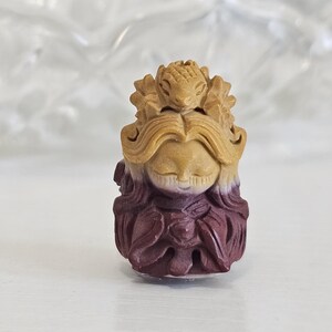 Op de afbeelding: Een klein uitgesneden beeldje met een figuur met gesloten ogen en vloeiend haar, bekroond met een hagedisachtig wezen. Het bovenste deel is lichtbruin en gaat over in een diepe, donkerpaarse kleur aan de basis, met gedetailleerde texturen.