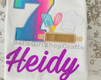 May include: White t-shirt with a rainbow ombre number 7, a chef's hat, a rolling pin, a whisk, and a spatula. The name "Heidi" is written in purple glitter below the design.