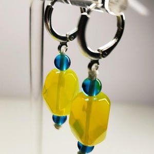 May include: A pair of silver-toned hoop earrings featuring dangling charms. Each charm consists of a large, faceted, translucent yellow bead positioned between two smaller, round, translucent blue beads on a thin cord.