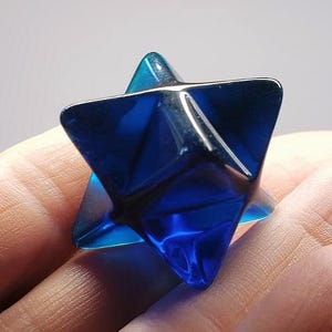 May include: A faceted, blue crystal Merkaba star. The geometric shape has six points, with a dark interior and reflective surfaces. The crystal is held in a hand, with a soft background.
