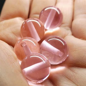 May include: Five translucent, round beads with a pinkish-orange hue are held in a hand. Each bead has a white, rod-like inclusion running through its centre. The beads are smooth and appear to be made of glass or a similar material.