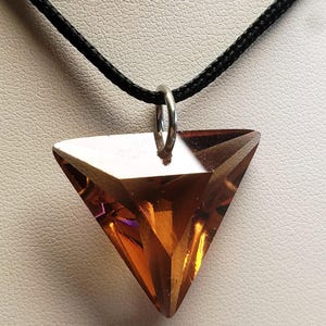 May include: A faceted, triangular-shaped amber-coloured pendant suspended from a thin black cord. The pendant features sharp, geometric edges and a polished, translucent surface that catches the light, attached to the cord by a silver-toned metal ring.