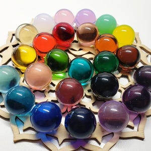 Può includere: Una collezione di biglie di vetro colorate in varie tonalità di rosso, arancione, giallo, verde, blu, viola e trasparente. Le biglie sono disposte su una base di legno decorativa, creando un'esposizione vibrante di colori e trame.