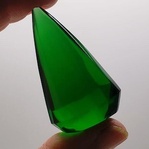 May include: A faceted, emerald-green gemstone with a pointed top and geometric sides. The gem is held between two fingers against a white background, showcasing its clarity and vibrant colour. The shape is reminiscent of a crystal.
