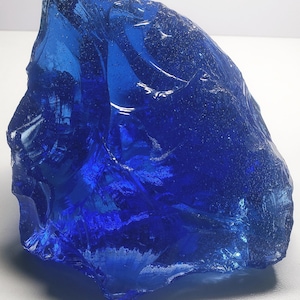 Puede incluir: Un gran trozo de vidrio azul profundo de forma irregular. El vidrio tiene una superficie rugosa y texturizada con muchas facetas y reflejos internos, creando un efecto brillante. La forma general es algo triangular, con un borde dentado.