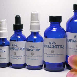 May include: An assortment of cobalt blue glass bottles with various tops, including droppers and sprayers. Sizes range from 1/2 oz to 8 oz, each labeled with white stickers. A tropical flower and greenery are in the foreground.