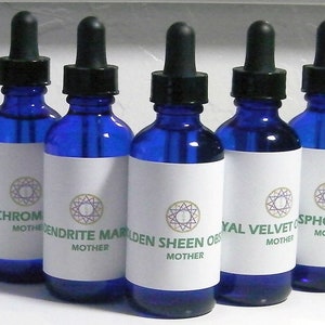 May include: A collection of blue glass dropper bottles with black caps and white labels. Each label features a gemstone name and the word "MOTHER" in green text. The bottles are arranged in a row, showcasing a variety of mineral-based products.