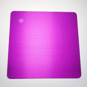 Puede incluir: Una alfombrilla de ratón cuadrada de color morado brillante con un acabado de metal cepillado. La alfombrilla de ratón tiene un pequeño logotipo en la esquina superior izquierda.