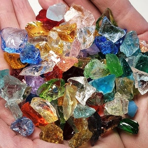 Puede incluir: Un conjunto de piezas de vidrio de colores, de forma irregular, que incluyen rojo, azul, verde, amarillo y transparente. Las piezas de vidrio se sostienen en la palma de una mano abierta, sobre un fondo neutro.
