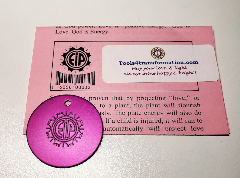 May include: A pink round metal disc with a black gear design and the letters "EIP" inside. The disc has a small hole at the top for hanging. The disc is next to a pink card with the text "Tools4transformation.com May your love & light always shine happy & bright!" and a barcode.