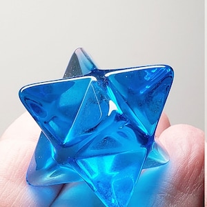 May include: A blue glass star-shaped ornament. The geometric form has sharp, triangular points and facets that reflect light. The object is held in a hand, against a plain background.