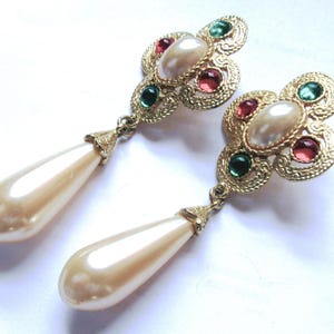 May include: A pair of gold-tone drop earrings featuring a textured clover-shaped top set with a central cream-colored faux pearl, flanked by small round green and pink stones. A long, smooth, cream-colored teardrop faux pearl hangs from each base.