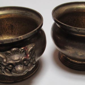 Op de afbeelding: Twee antieke messing wierookbranders met drakenkop handvatten. De branders zijn rond en hebben een getextureerd oppervlak.