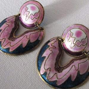 May include: A pair of vintage earrings with a gold-tone metal finish. The earrings feature a circular top and a teardrop-shaped bottom, with pink, blue, and white enamel detailing. The design includes a wavy pattern and a small pink circle.