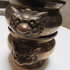 Puede incluir: Dos recipientes de latón apilados con un diseño decorativo de cabeza de león. Los recipientes son de color dorado oscuro y tienen un acabado liso y pulido.