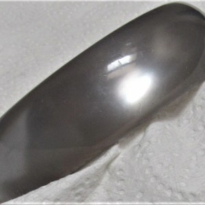 Puede incluir: Un brazalete liso de color gris hecho de plástico. El brazalete es brillante y tiene una forma redondeada.