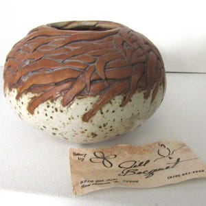 May include: A handcrafted ceramic vase with a round shape and a wide opening. The upper portion features a brown leaf design, while the lower half is speckled white. A small tag with the name "Jill Becquet" is visible.