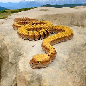 May include: A yellow and brown snake with a scaly texture is coiled on a light gray rock. The snake has a distinctive head with a yellow eye.