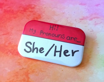 Può includere: Bottone rosso e bianco con il testo "Hi! My Pronouns are... She/Her" su uno sfondo ad acquerello.