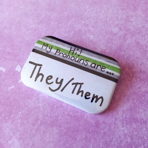 May include: A white button with a green, white and black striped border. The text on the button reads "Hi! My Pronouns are... They/Them".