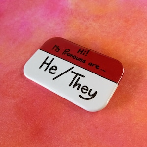 Puede incluir: Una etiqueta de nombre roja y blanca con el texto "Hi! My Pronouns are... He/They" escrito con un marcador negro.