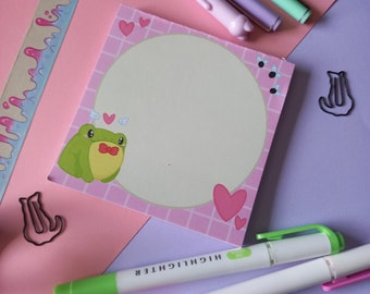 May include: A pink and white notepad with a grid pattern and a cartoon frog wearing a bow tie. The notepad has a circle in the center with a white background. The notepad is surrounded by pink hearts and flies. The notepad is on a pink and purple surface with two highlighters.