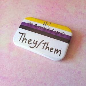 May include: A white button with yellow, purple and black stripes. The text on the button reads "Hi! My pronouns are They/Them".