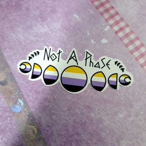 Puede incluir: Una pegatina con cinco lunas en diferentes fases, cada una con los colores de la bandera del orgullo no binario. El texto "Not A Phase" está escrito sobre las lunas.