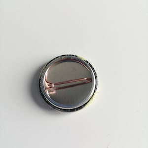 Puede incluir: Un bot&oacute;n redondo con una parte trasera de metal plateado y un cierre de alfiler de seguridad. El borde del bot&oacute;n es negro con un dise&ntilde;o amarillo y verde. El bot&oacute;n est&aacute; sobre un fondo blanco.