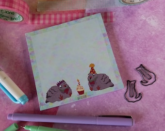 May include: A light blue notepad with a whimsical illustration of two birds wearing party hats and a cupcake with a candle. The notepad has a white border and a light blue background.